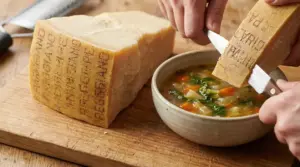 Mani che aggiungono una scorza di parmigiano a una zuppa di verdure accanto a un pezzo di formaggio