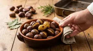 Ciotola di castagne arrosto su tavolo di legno, con una mano che la serve e teglia sullo sfondo